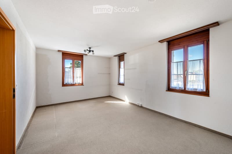 Empty room with white walls, carpeted floor, and wooden-framed windows with sheer curtains. Features a wooden door on the left and a ceiling light fixture.