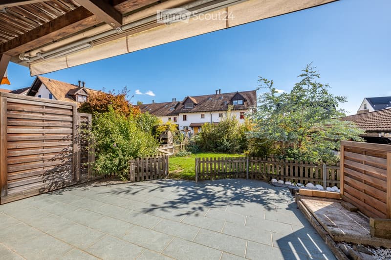 A well-maintained garden area with a paved patio, wooden fences, and surrounding greenery, featuring a clear blue sky and neighboring houses in the background.