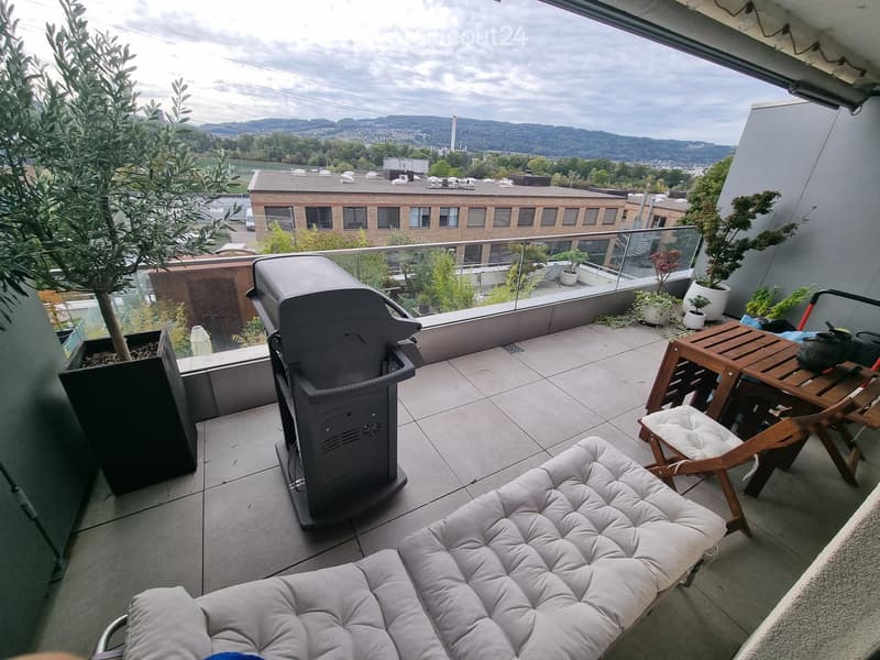 Balcony with a grey sofa, wooden table, chairs, grill, and potted plants, offering a scenic city view.