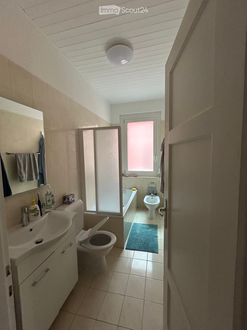 Bathroom with a sink, toilet, bathtub, mirror, and a shower stall. The room features tiled walls and floor, a window with frosted glass, and a small blue rug in the shower area.