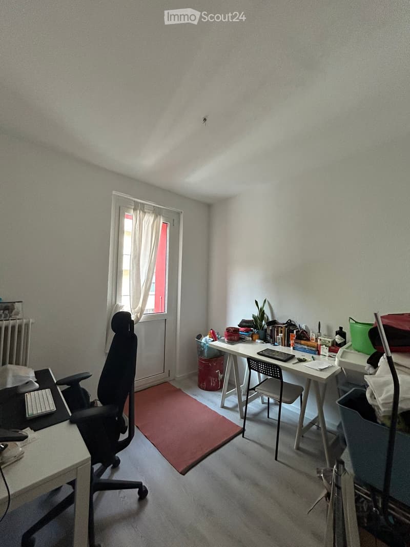 A furnished room with a desk, chair, laptop, keyboard, and a small plant on a side table. The room has a window with a curtain and a radiator below it.