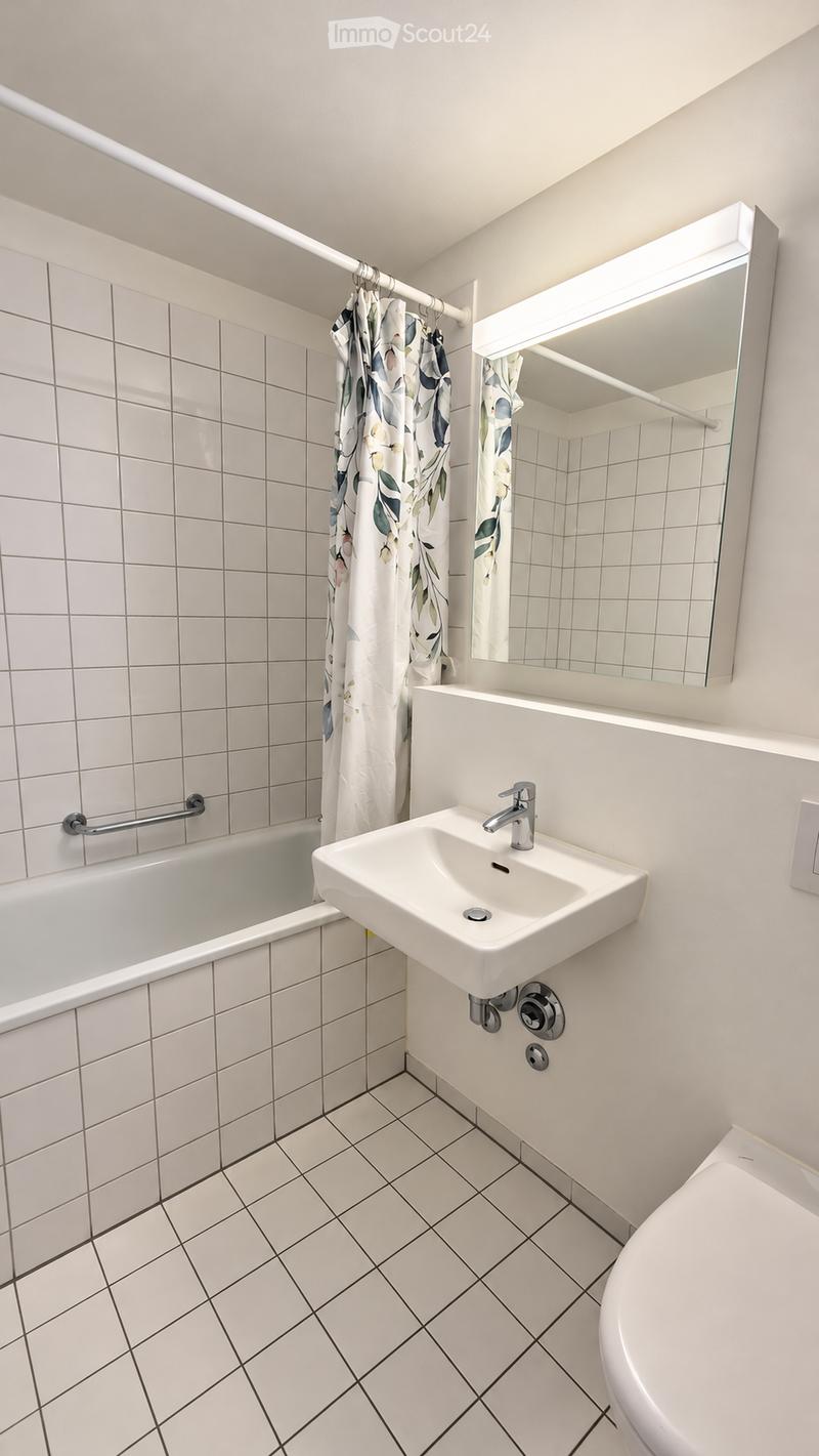 Modern bathroom with white tiled walls and floor, featuring a bathtub, sink, toilet, and a large mirror with built-in lighting. A floral-patterned shower curtain is visible.
