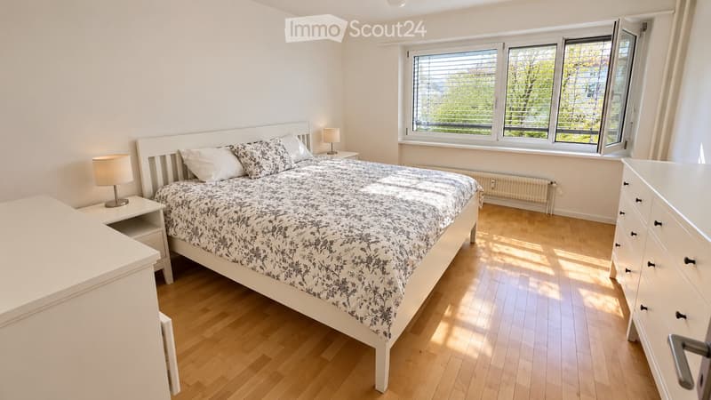 Bright bedroom with wooden flooring, large window with blinds, white bed with patterned bedding, nightstands with lamps, and a dresser.