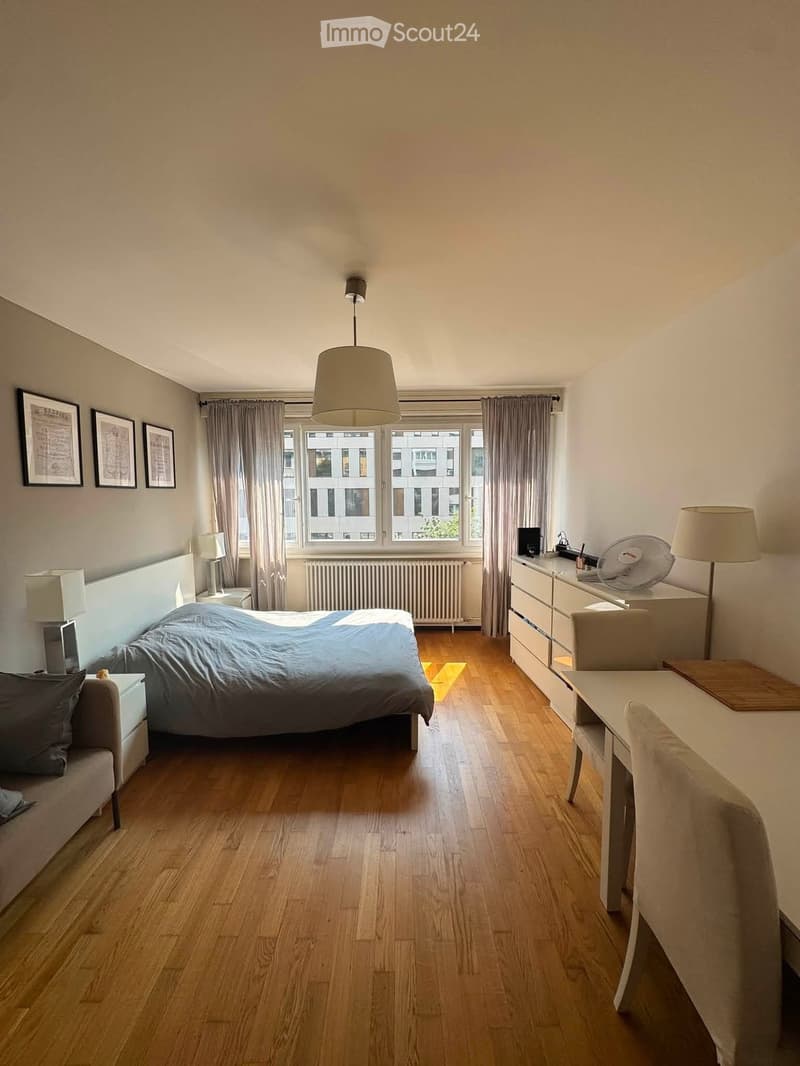 A well-lit bedroom with wooden flooring, a large bed with a blue duvet, a nightstand with a lamp, a desk with a chair, and a fan on top of a dresser. The room features large windows with curtains and several picture frames on the wall.
