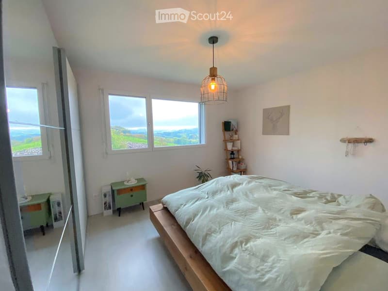Bright bedroom with a wooden bed, nightstand, hanging lamp, shelf, and scenic view through large windows.