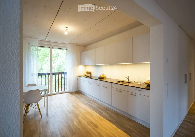 Modern kitchen with white cabinets, wooden flooring, and a small dining area. Features a sink, faucet, and a sliding glass door leading to a balcony.
