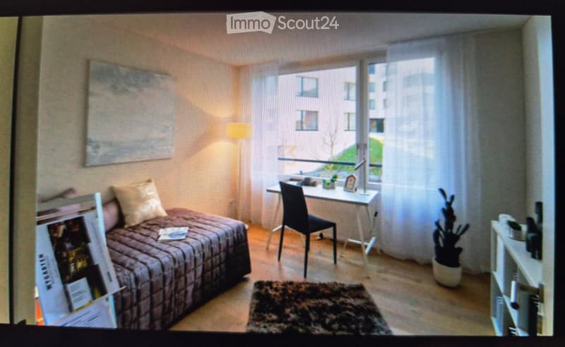 Cozy bedroom with a bed, desk, chair, lamp, and plant near the window. The room features a large window with a view of the city and a mirror on the wall.