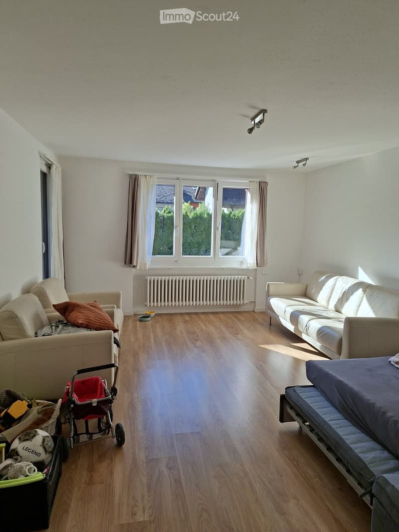 Living room with light-colored wooden flooring, large windows with curtains, a sofa, armchair, radiator, baby stroller, and a mattress on the floor.