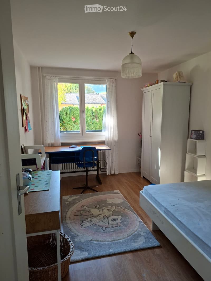 Well-lit bedroom with wooden flooring, a bed, desk, chair, wardrobe, and a round rug with a cartoon design.