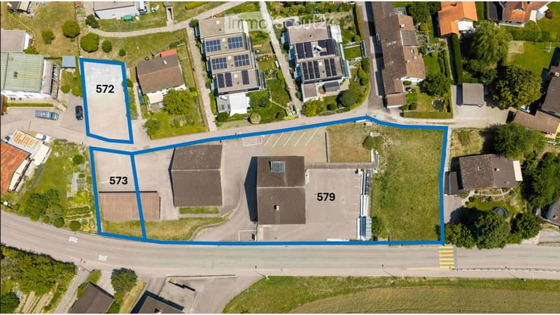 Aerial view of a residential area showing multiple buildings, parking lots, and surrounding greenery. Three plots are highlighted with blue outlines and labeled with numbers (572, 573, 579).