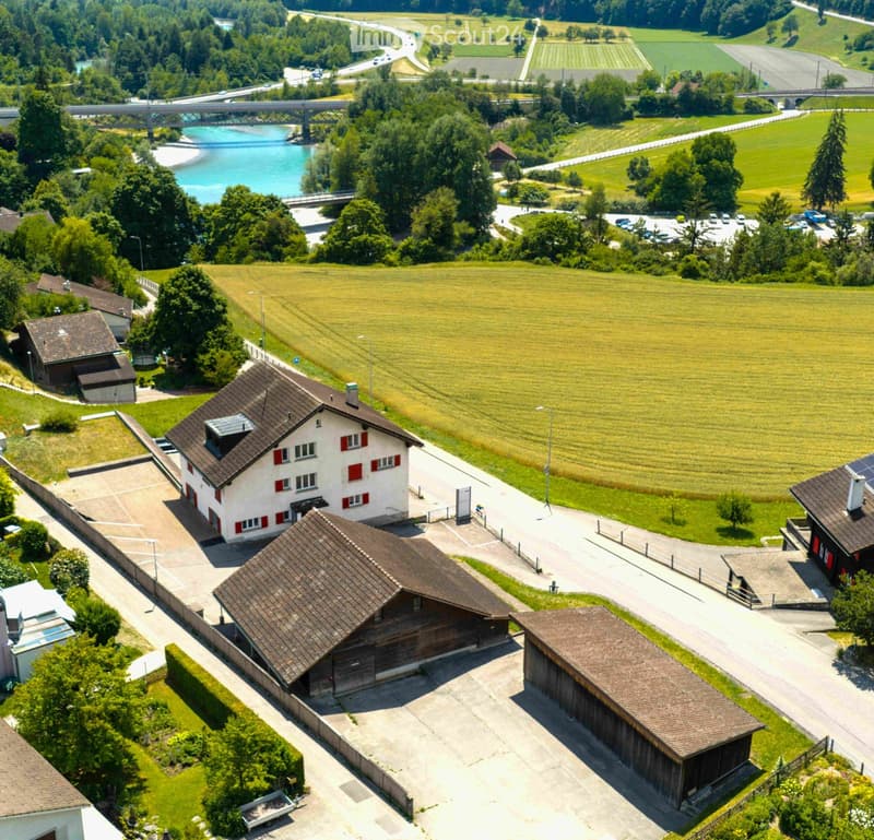 Aerial view of a rural residential area with multiple houses, a parking lot, and surrounding green fields and a river.