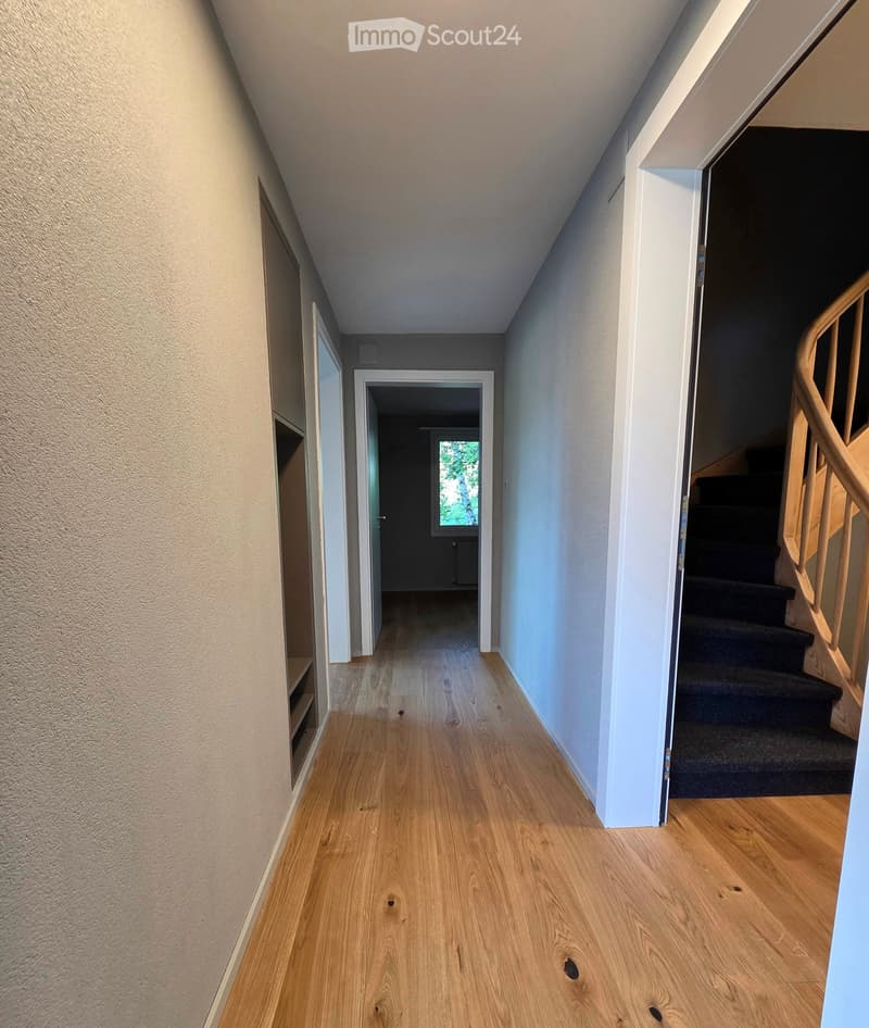 Bright hallway with wooden flooring, white walls, and a staircase leading upwards. The hallway has a door on the left and a window at the end, allowing natural light to enter.