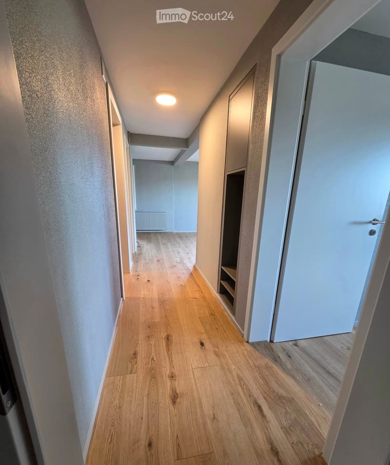 A modern hallway with light wooden flooring, white walls, and recessed lighting. Doors lead to other rooms, and a small built-in shelf is visible on the right side.