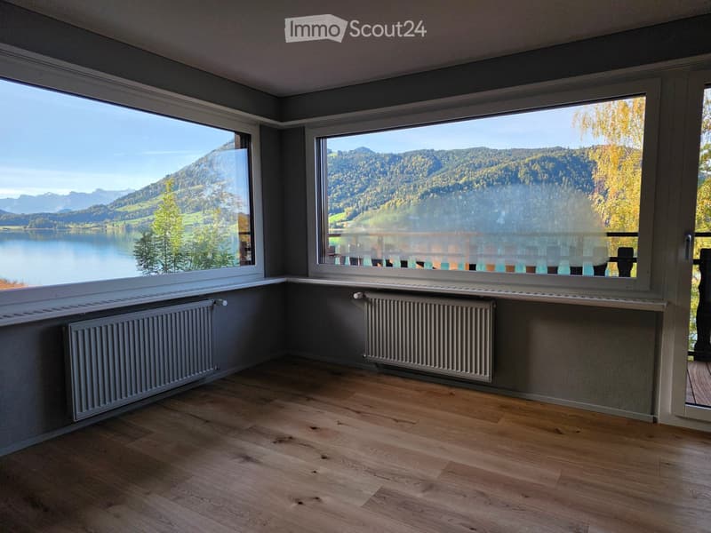 Empty room with wooden flooring, large windows offering a scenic view of a lake and mountains, and radiators along the walls.