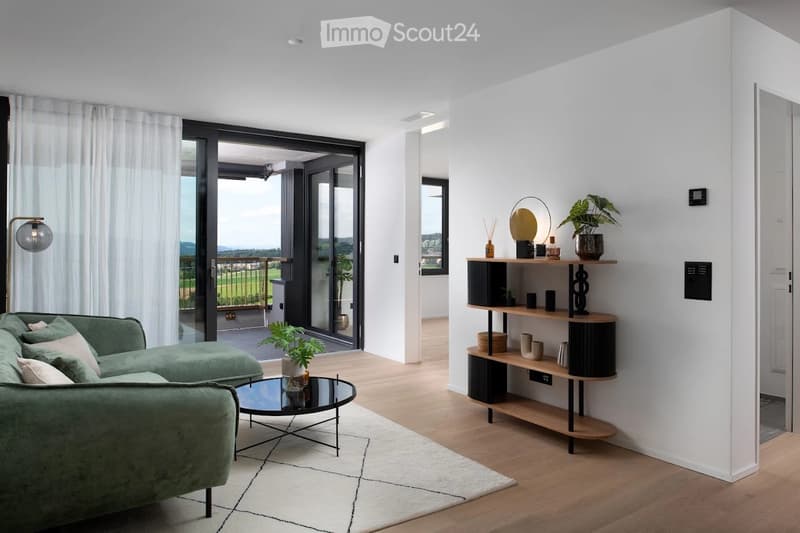Modern living room with a green sectional sofa, glass coffee table, wooden shelf with decorative items, and large windows offering a scenic view.