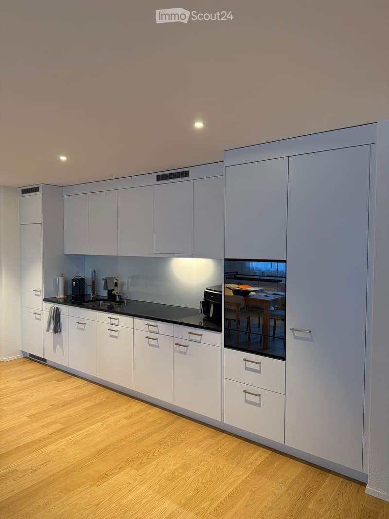 Modern kitchen with white cabinets, black countertops, and a reflective backsplash. Visible items include a sink, faucet, coffee maker, and oven. A small dining area with a table and chairs is visible through the backsplash.