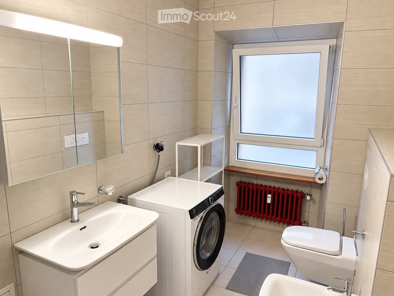 Modern bathroom with a white sink, toilet, washing machine, and a large mirror above the sink. The room features light wooden tiles and a red radiator under the window.
