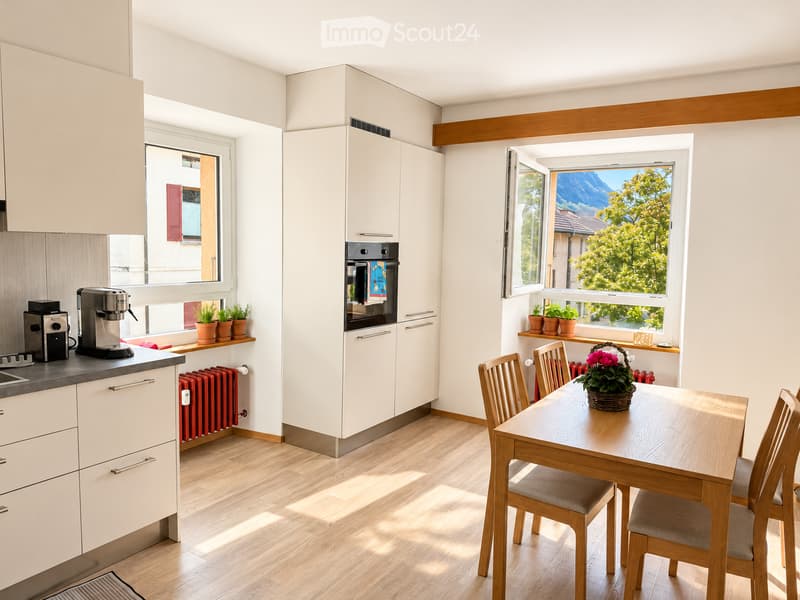 Bright kitchen with white cabinets, wooden floor, coffee maker, oven, dining table with chairs, and potted plants on windowsills.