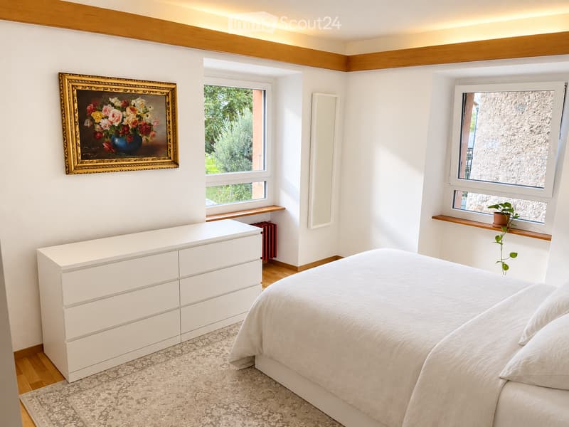 Modern bedroom with a large bed, white dresser, framed painting, small plant on windowsill, and radiator.