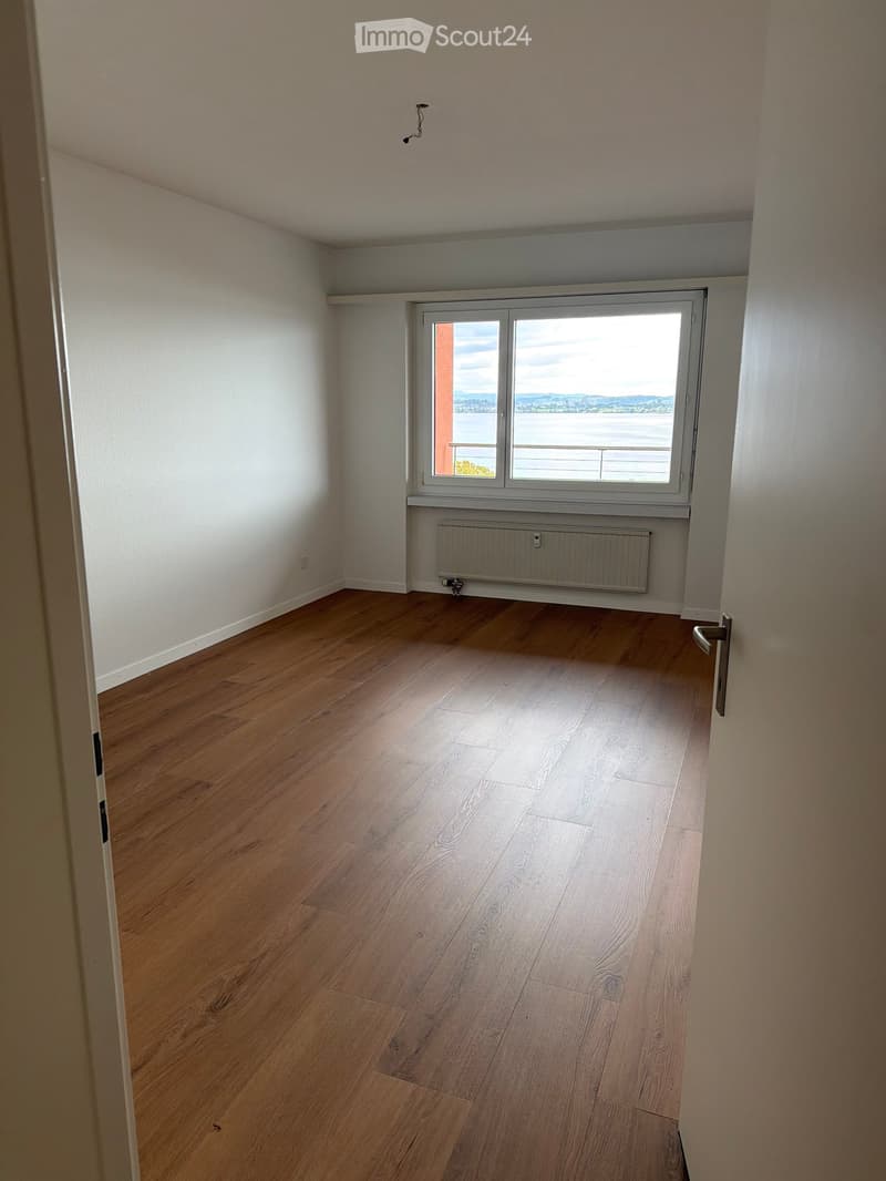 Empty room with wooden flooring, white walls, and a large window offering a scenic view.