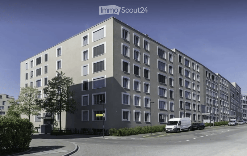 A modern multi-story apartment building with a grey facade, numerous windows, and a clean, contemporary design. The building is surrounded by a small landscaped area with trees and shrubs, and a few vehicles are parked along the street in front of it.