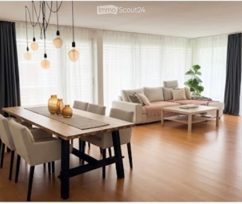 Bright living and dining area with wooden flooring, large windows, a sofa, coffee table, dining table with chairs, and decorative vases.