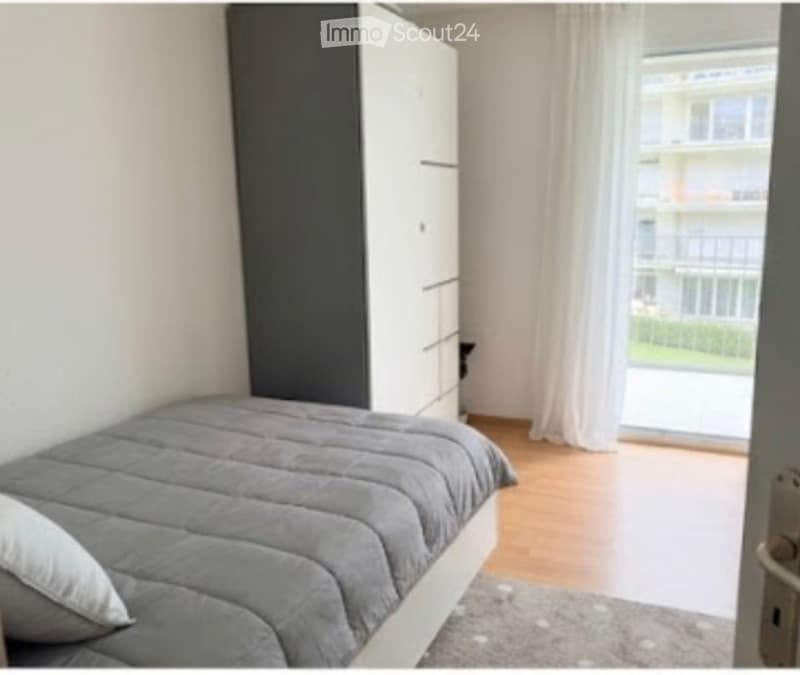Modern bedroom with a grey duvet-covered bed, white wardrobe, and large window with white curtains leading to a balcony.