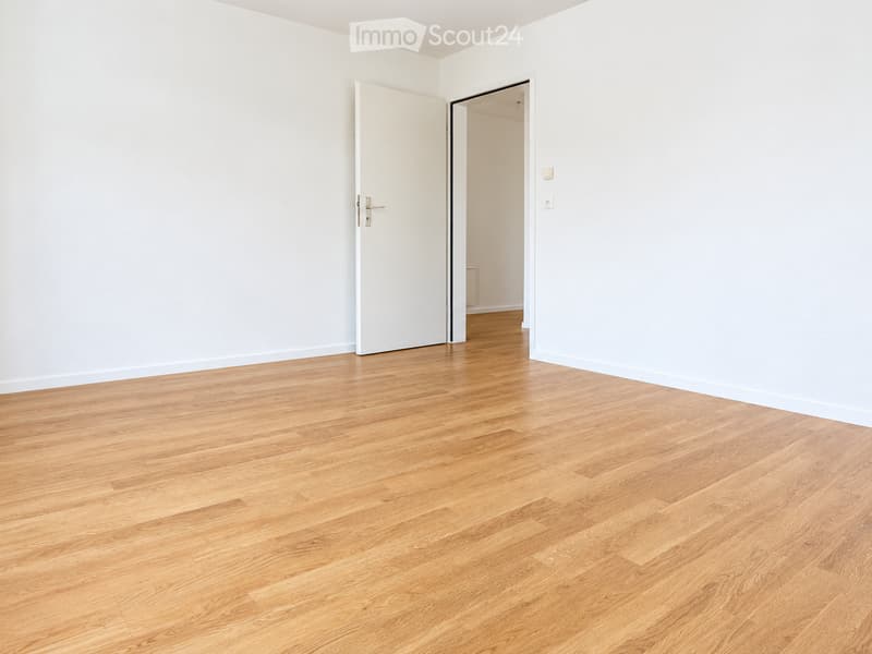 Empty room with light wooden flooring and white walls, featuring an open doorway leading to another room.