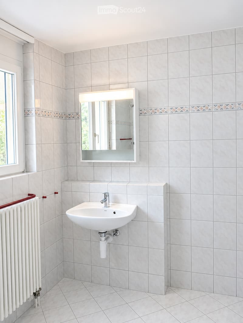 Modern bathroom with beige tiled walls and floor, white sink with chrome faucet, illuminated mirror above, and a white radiator on the left side.