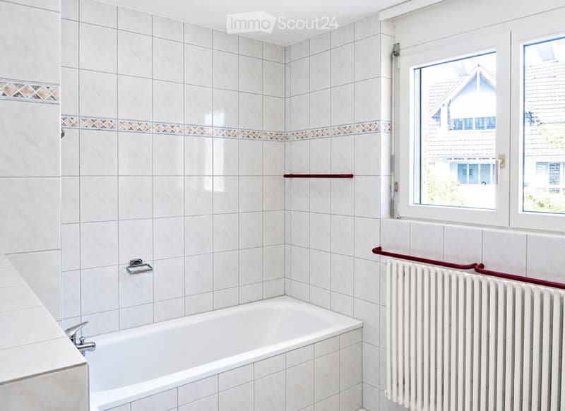 Bathroom with white tiled walls and floor, a bathtub, a radiator, and a handrail near the window.