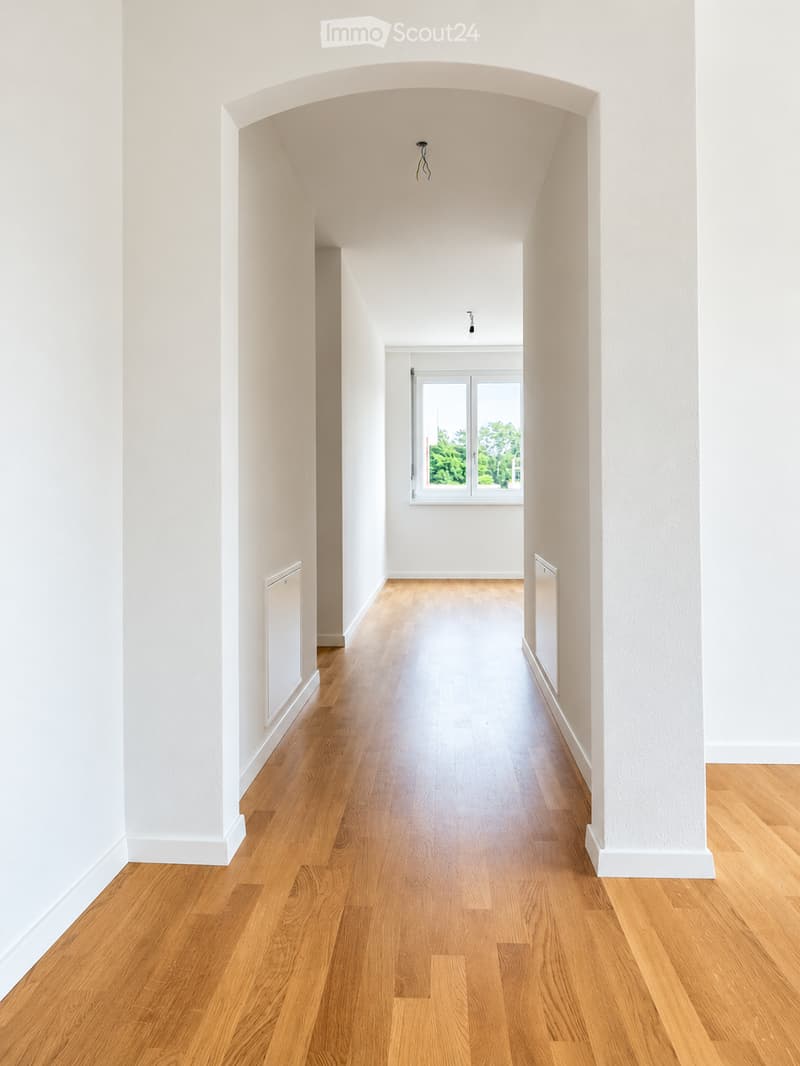 Empty hallway with light wooden flooring, white walls, and a large window at the end showing greenery outside.