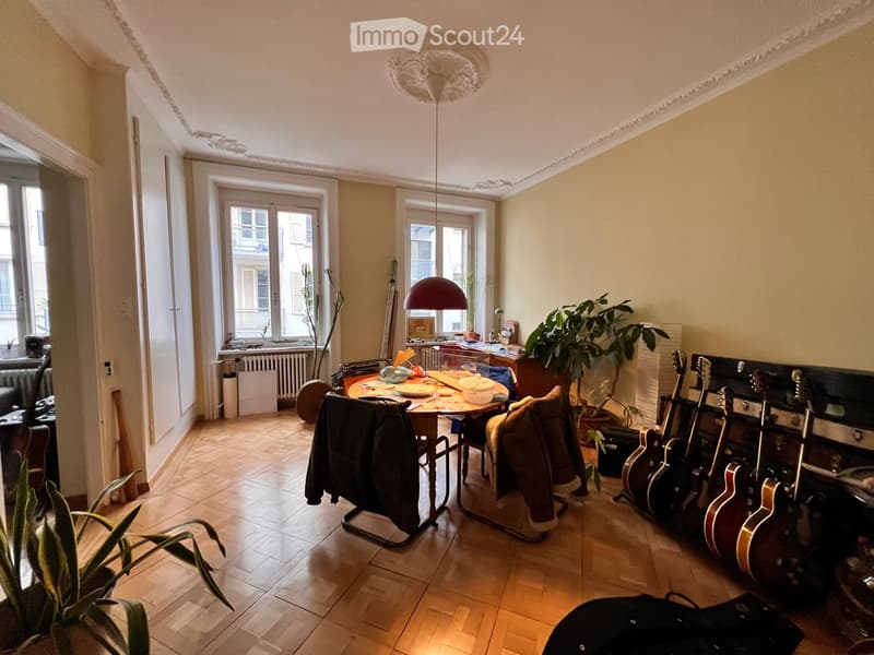 A well-lit room with wooden flooring, a dining table with chairs, several guitars leaning against the wall, and potted plants. The room features large windows and a cozy ambiance.