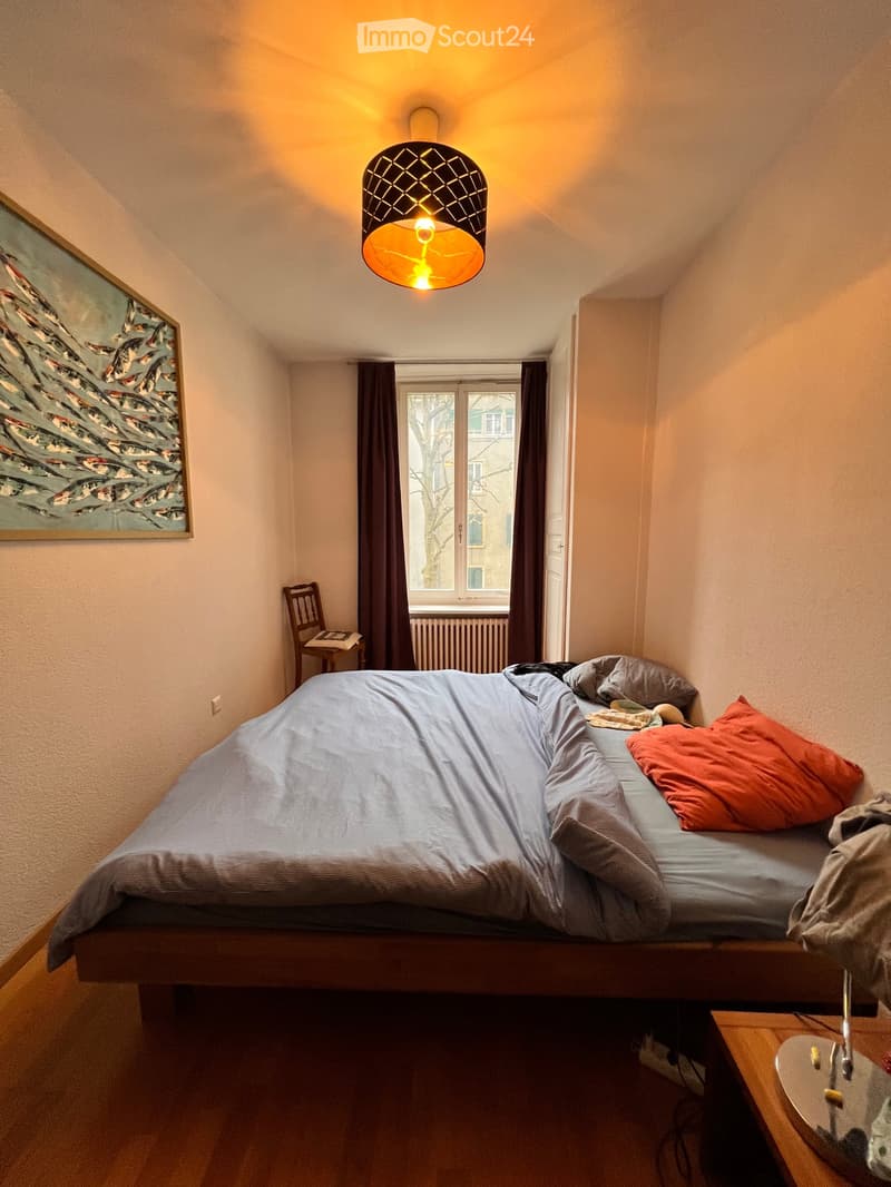 Cozy bedroom with a double bed, wooden chair, small table, and a painting on the wall. The room features warm lighting from a hanging lamp and has a window with dark curtains.