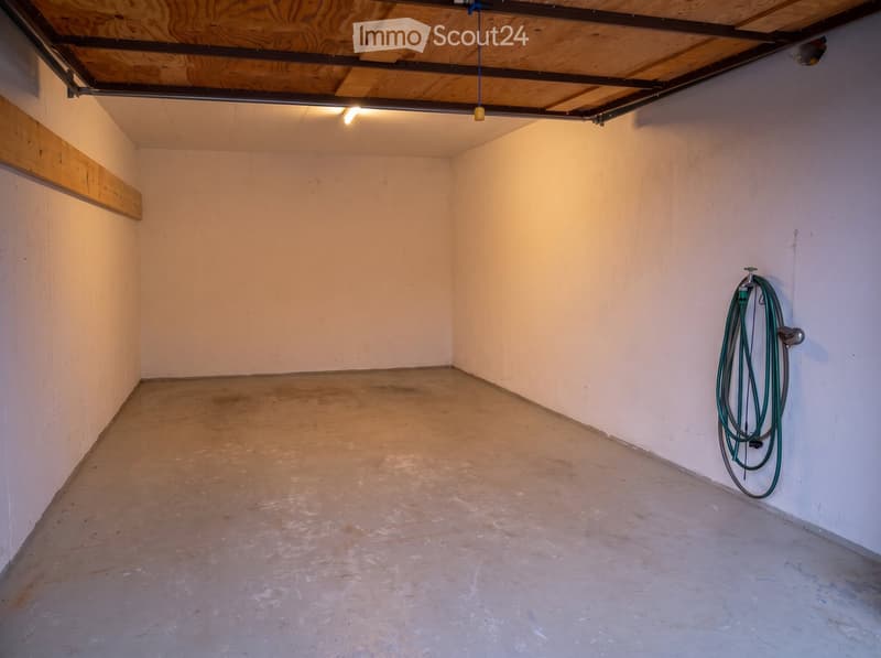 Empty room with concrete floor, exposed wooden beams, and a coiled green hose on the wall.