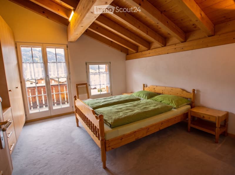 Cozy attic bedroom with wooden beams, large windows offering a scenic mountain view, a double bed with green bedding, a nightstand, and a wooden chair.