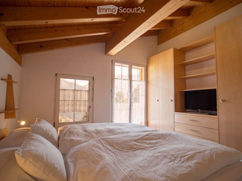 Cozy bedroom with a large bed, pillows, a wardrobe, shelves, and a television. The room features wooden beams on the ceiling and large windows with curtains.