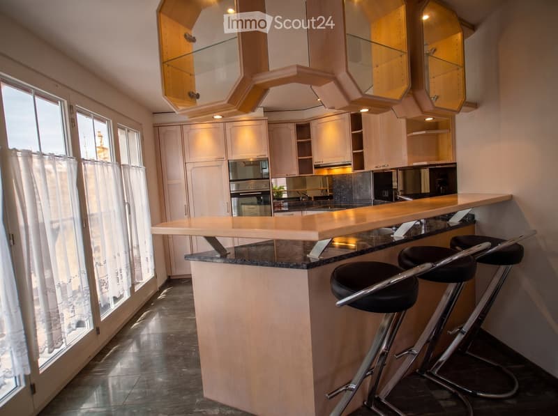 Modern kitchen with white cabinets, a central bar counter, and two black bar stools. Features large windows with white curtains and a sleek, contemporary design.