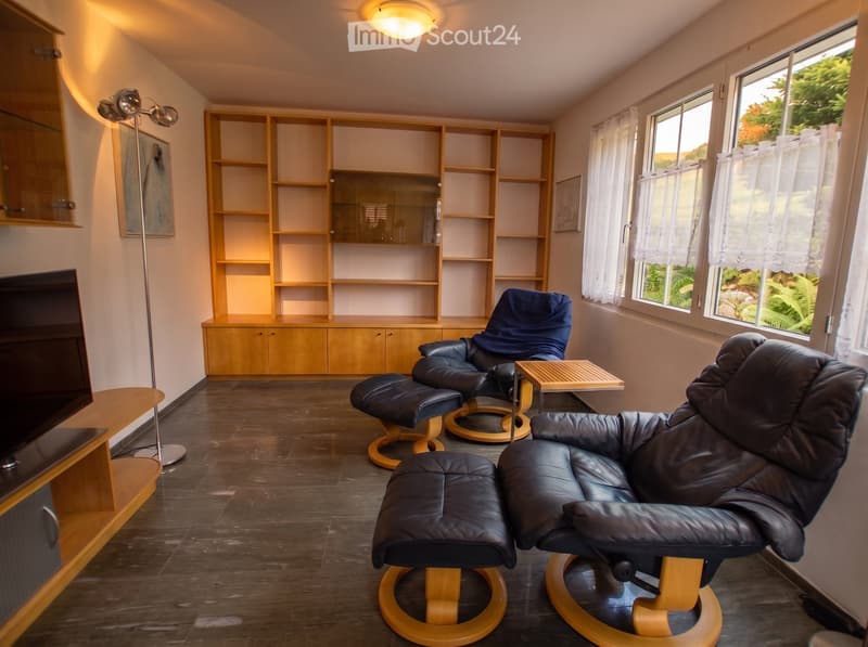 Modern living room with dark wooden flooring, large windows with white curtains, two leather chairs with matching ottomans, a small wooden side table, built-in wooden shelving unit, and a TV stand with a flat-screen TV.