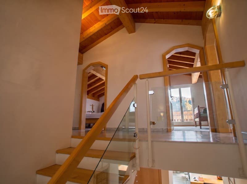 Staircase with wooden steps and glass railing, leading to a bedroom area with visible bed and furniture.