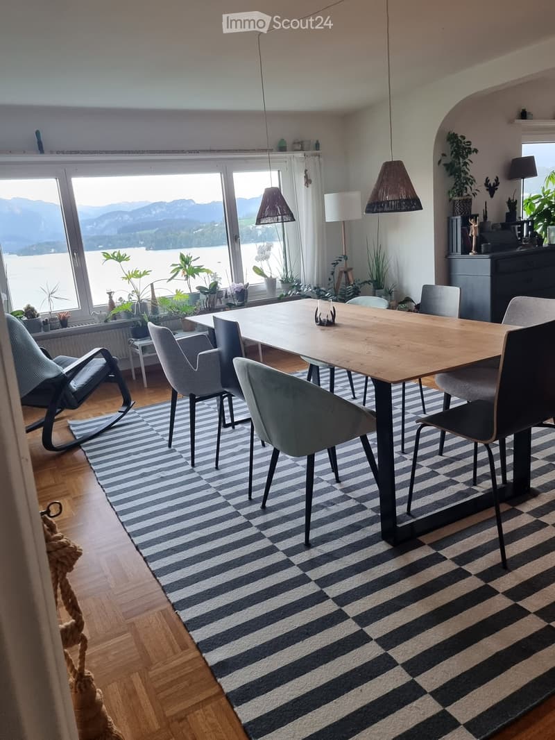 Bright dining area with a wooden table, six chairs, a striped rug, and large windows offering a scenic lake and mountain view. Includes a hanging lamp, side table, and decorative plants.