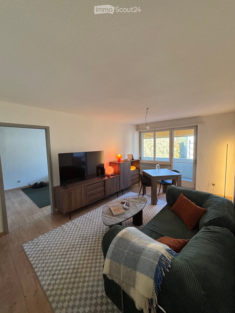 Living room with a green sofa, coffee table, television, sideboard, dining table, and chairs. The room features wooden flooring, a patterned rug, and large windows with a view.