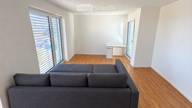 Bright living room with wooden flooring, large windows with blinds, grey sectional sofa, small desk, and chair.