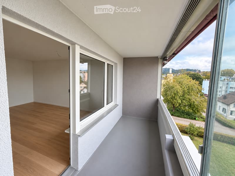 Balcony with wooden floor, large windows, and a view of greenery and buildings.