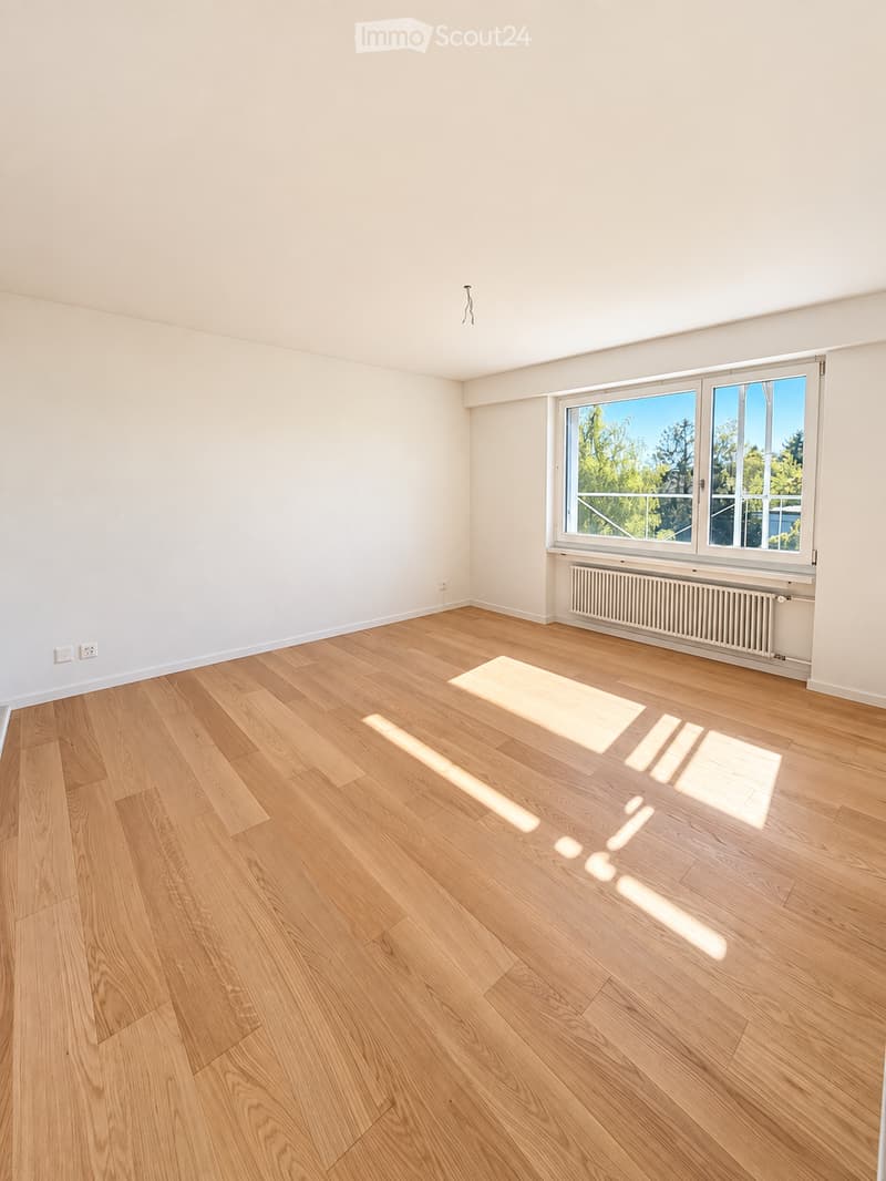 Empty room with light wooden flooring, white walls, and large windows allowing natural light to enter. The room features a radiator below the windows and electrical outlets on the walls.