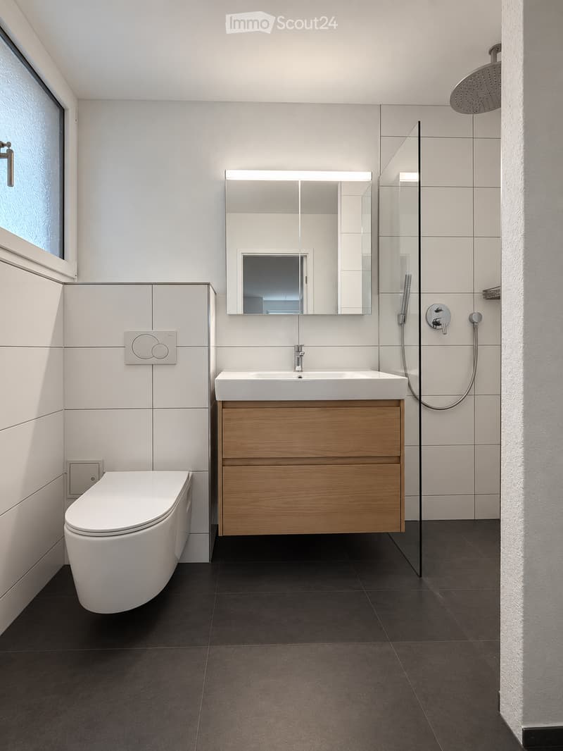 Modern bathroom with white tiles, dark floor, wooden vanity, sink, toilet, mirror, and glass-enclosed shower area.