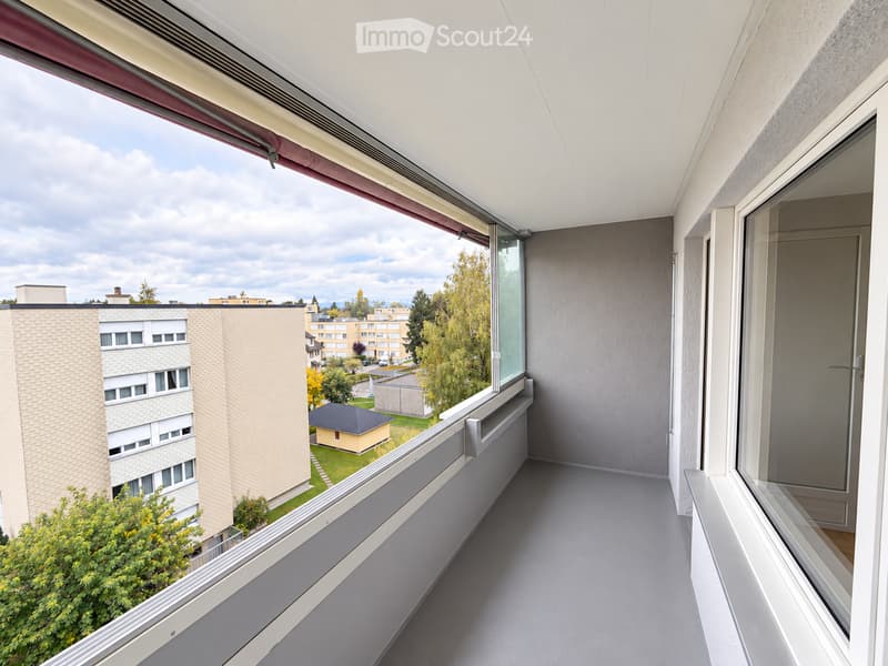 Modern balcony with grey flooring, large windows offering a view of surrounding buildings and greenery.