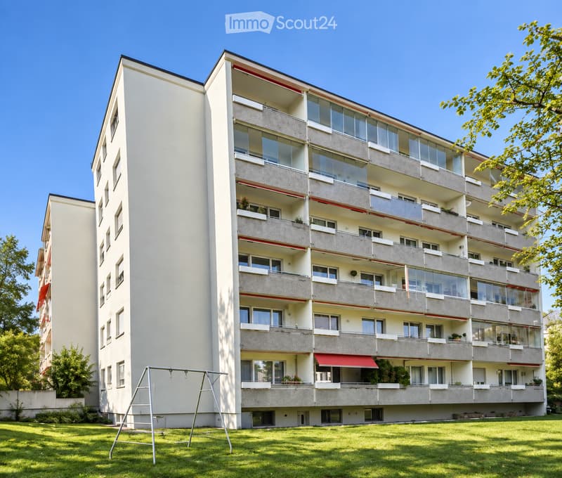 Multi-story residential building with balconies, large windows, and a playground in the foreground.