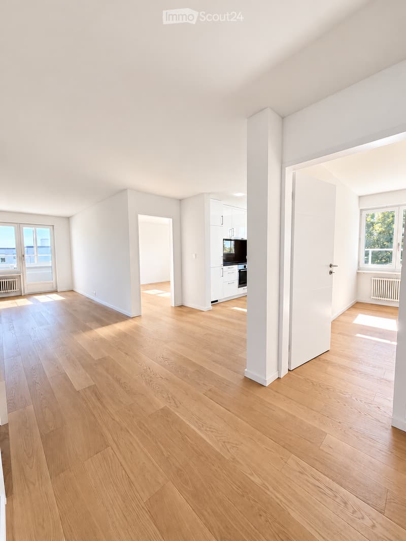Bright, empty room with light wooden flooring, white walls, and large windows allowing natural light. The space features a central column and an open doorway leading to another area with white cabinetry and appliances.