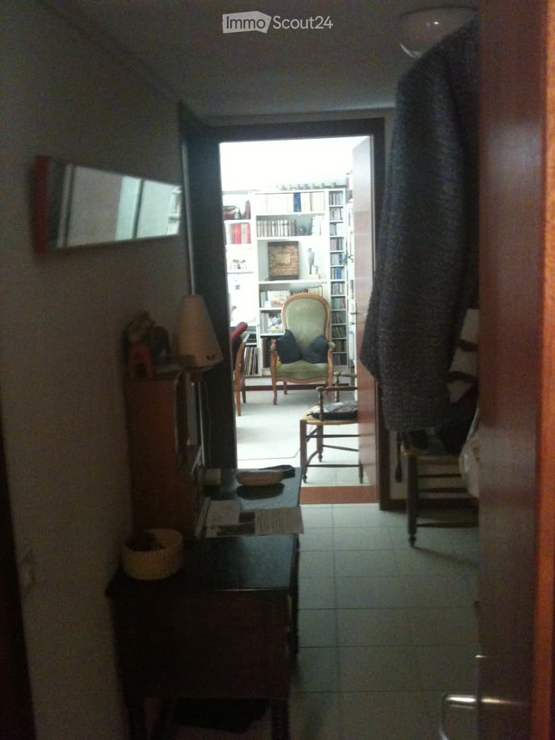 A narrow hallway with tiled flooring, leading to a room with bookshelves, a green armchair, and a small table. The hallway features a small side table with a lamp and a basket, and a mirror on the wall.