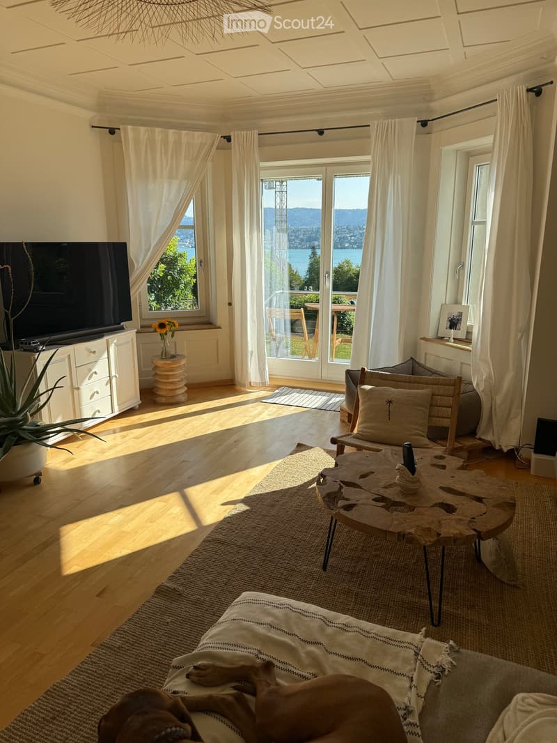 Bright living room with wooden floors, large windows offering a scenic view, a sofa with a dog resting on it, a coffee table, a side table with a flower vase, a television, and a cabinet.